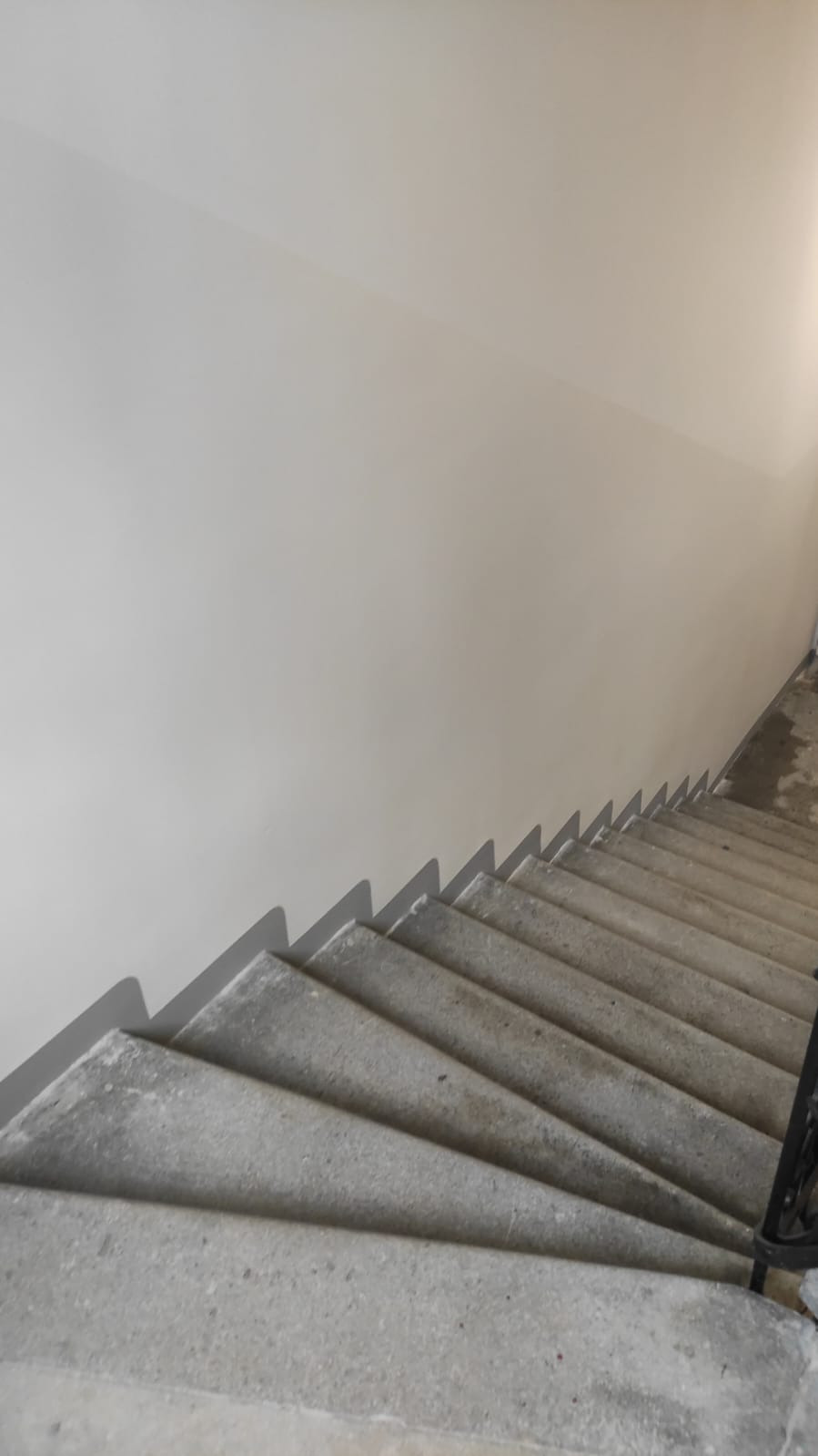 Świeżo pomalowana ściana klatki schodowej w jasnym, neutralnym odcieniu szarości, widoczna z góry. Betonowe schody z czarną balustradą.