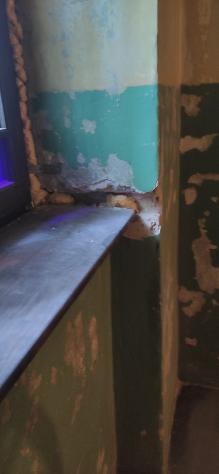 Narożnik pokoju z oknem, ściany z odpryskującą farbą w odcieniach zieleni i beżu, ukazujące surowy stan przed remontem. Widoczne uszkodzenia i brak wykończenia.