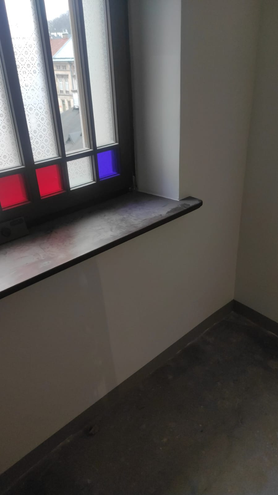Wykończone wnętrze: świeżo pomalowana ściana w jasnym odcieniu, ciemny parapet z kolorowymi szybkami w oknie. Widok na budynek z zewnątrz.