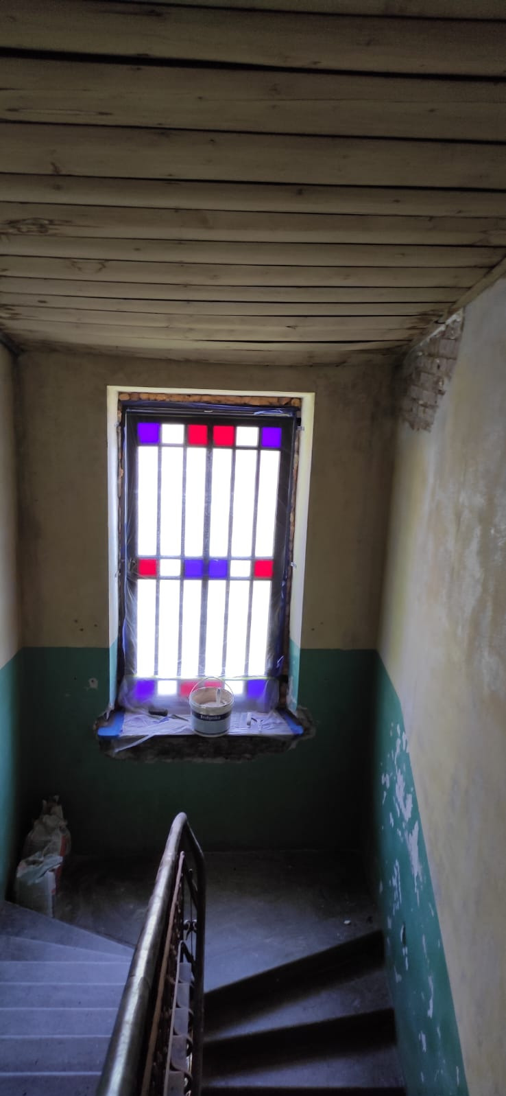 Klatka schodowa w trakcie renowacji: ściany częściowo pomalowane na zielono i żółto, okno z kolorowymi szybkami, drewniany sufit, widoczny kubełek z farbą.