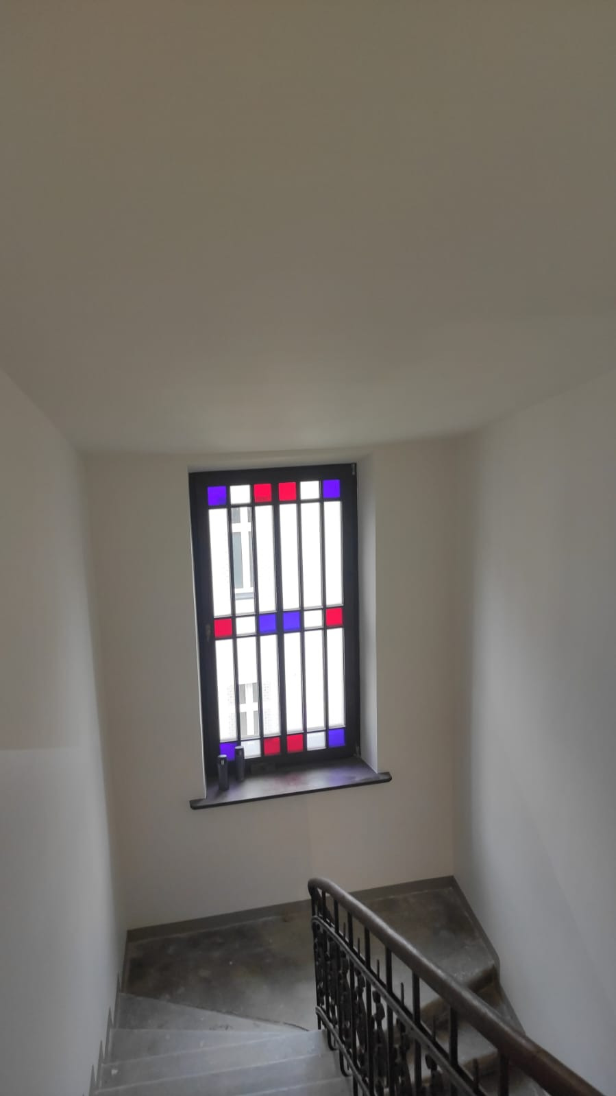 Klatka schodowa z oknem w kolorowe kraty. Świeżo pomalowane ściany na biało. Balustrada schodów w ciemnym kolorze. Widok z góry na dół.