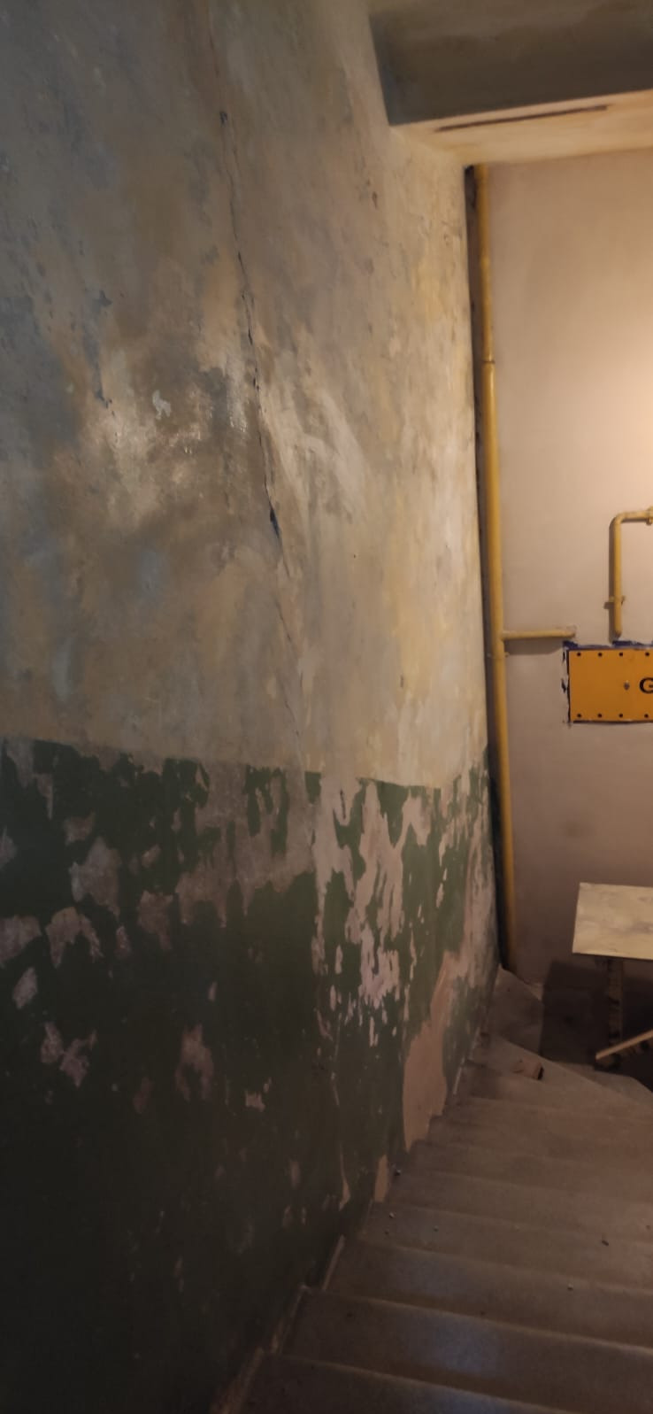Zniszczona ściana z widocznymi pęknięciami i odpadającą farbą, fragment schodów i żółta rura przy ścianie w tle. Widoczne ślady wcześniejszych prac remontowych.
