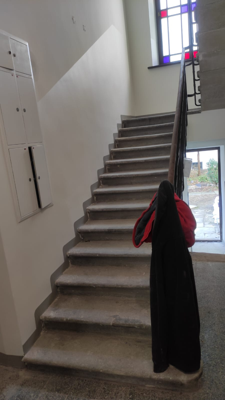 Schody z szarego kamienia w klatce schodowej, widoczne szafki na ścianie i okno z kolorowymi szybami. Na poręczy wisi czarny płaszcz z czerwoną podszewką.