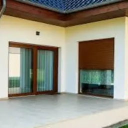 Nowoczesny taras z dużymi przesuwnymi drzwiami szklanymi i oknami z brązowymi roletami zewnętrznymi, widok z zewnątrz, jasna elewacja domu.