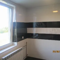 Wykończona łazienka z czarno-białymi płytkami z motywem róży, widok na okno z grzejnikiem pod spodem, data wykonania zdjęcia widoczna w prawym dolnym rogu.