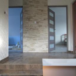 Osoba prywatna - Wykończone wnętrze z kamienną okładziną ściany, brązowymi płytkami podłogowymi i częściowo widocznymi drzwiami wewnętrznymi z przeszkleniami.