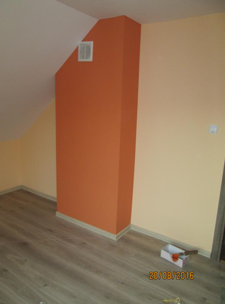 Świeżo pomalowane na pomarańczowo naroże w pokoju w Jodłowej, z widocznymi panelami podłogowymi i narzędziami malarskimi w kartonowym pudełku.