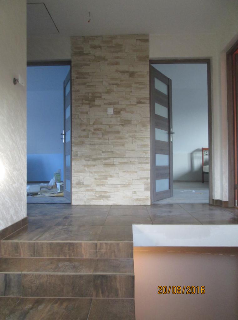 Wykończone wnętrze z kamienną okładziną ściany, brązowymi płytkami podłogowymi i częściowo widocznymi drzwiami wewnętrznymi z przeszkleniami.