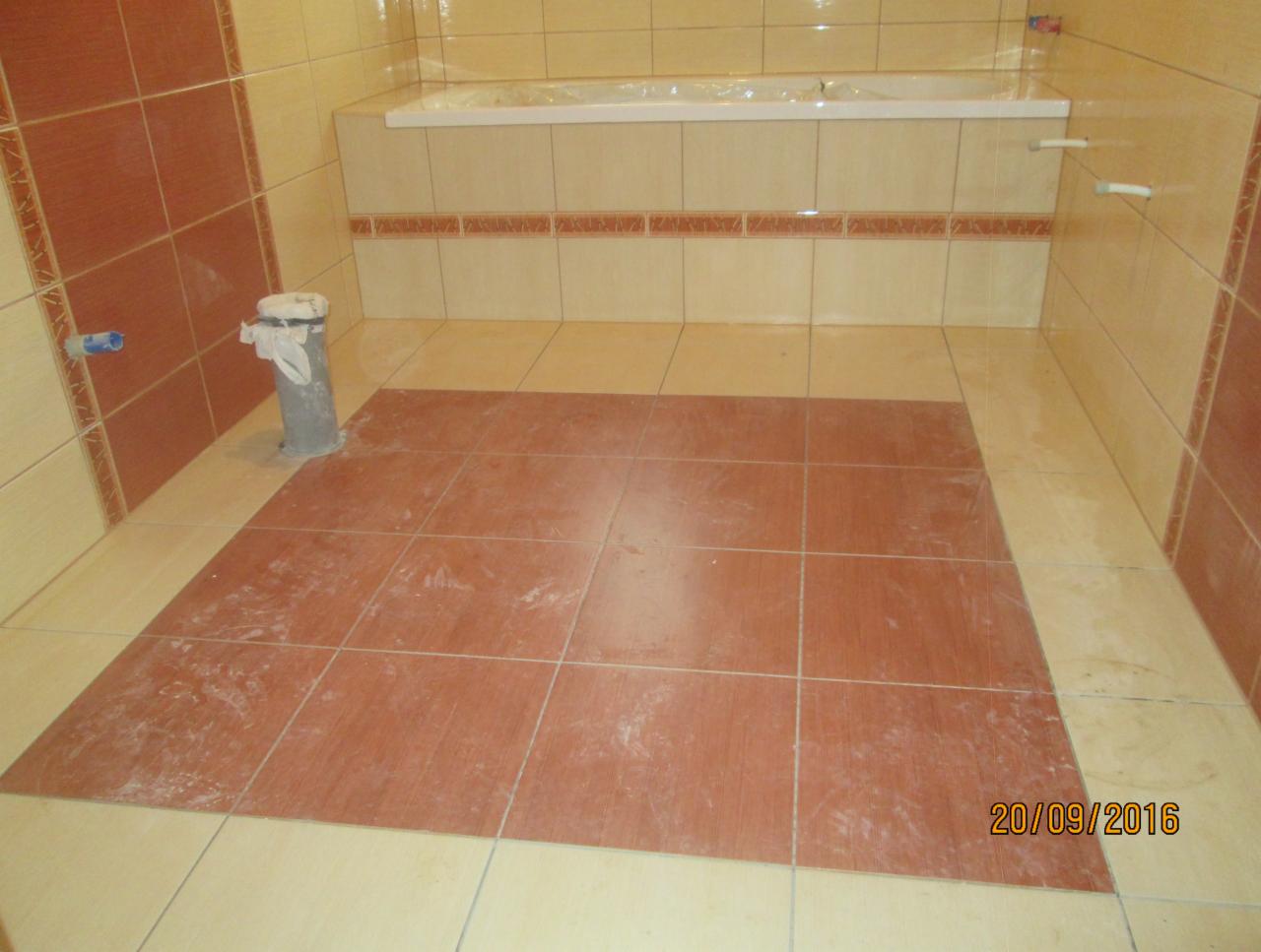 Częściowo ułożone, kwadratowe, terakotowe płytki podłogowe w łazience z beżowymi ścianami i wanną, widoczny fragment instalacji kanalizacyjnej.