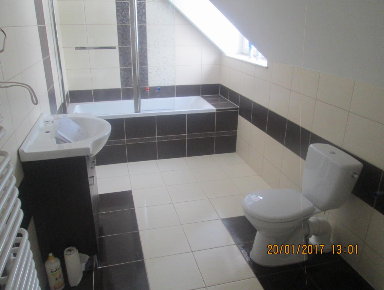 Wykończona łazienka na poddaszu z biało-czarnymi płytkami, umywalką na szafce, toaletą i wanną z parawanem.