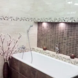 Elegancko wykończona łazienka w Gdańsku z wanną, beżowymi płytkami z dekoracyjnym motywem roślinnym, mozaiką w odcieniach brązu we wnęce oświetlonej punktowo oraz wazonem z kwitnącymi gałązkami.