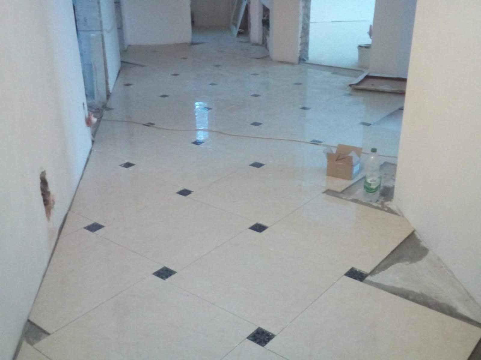 Układanie jasnych płytek podłogowych z ciemnymi wstawkami w korytarzu, widoczne niedokończone fragmenty i butelka wody mineralnej.