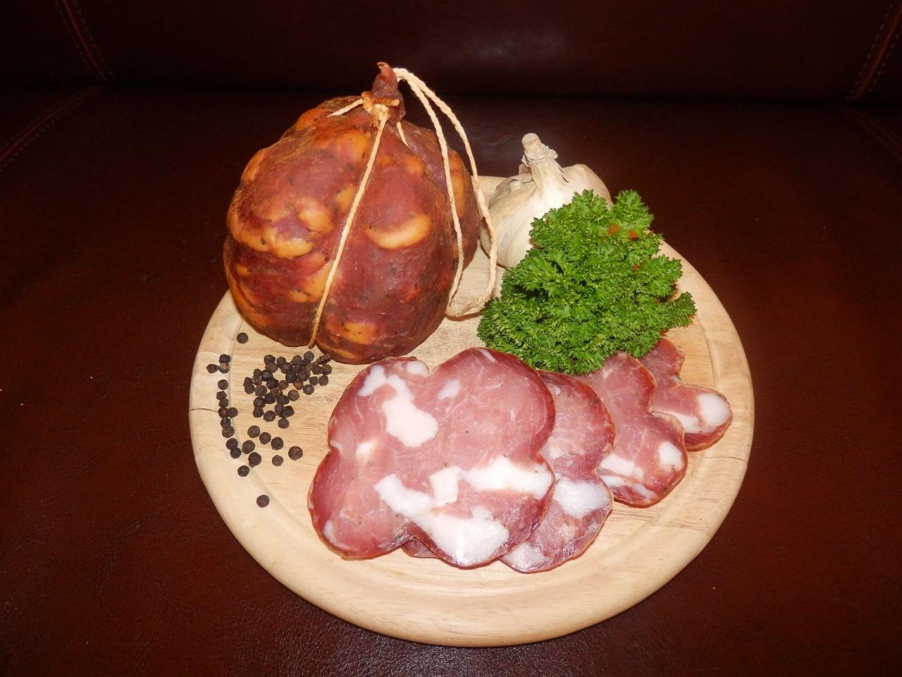 Okrągła deska z trzema plasterkami wędliny o nieregularnych kształtach, obok główka czosnku, natka pietruszki, kilka ziaren pieprzu i cała wędlina związana sznurkiem, na ciemnobrązowym tle.