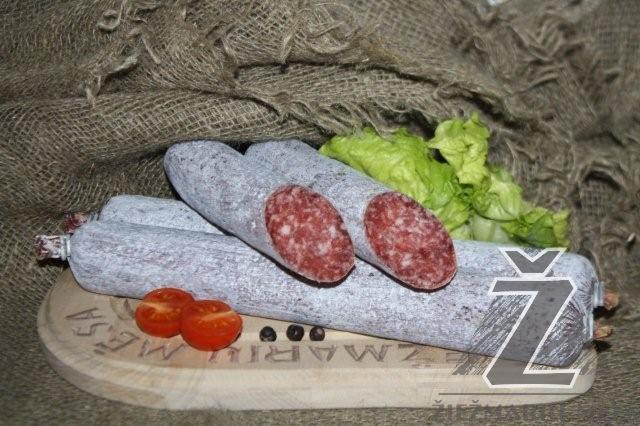 Suszona kiełbasa na drewnianej desce z wygrawerowanym napisem, udekorowana pomidorkami cherry, ziarnami pieprzu i liśćmi sałaty na tle jutowego materiału.