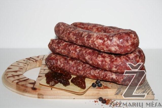 Trzy surowe kiełbasy ułożone jedna na drugiej na drewnianej desce w N. Kietaviskiu k., obok suszone pomidory i jagody, całość na białym tle.