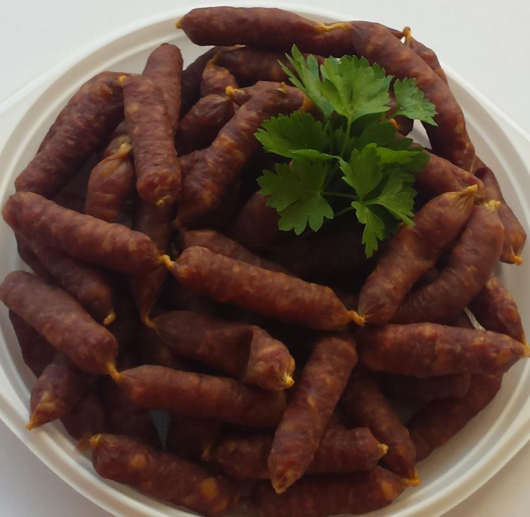 Kiełbaski kabanosy ułożone na białym talerzu, udekorowane gałązką zielonej pietruszki, widok z góry.