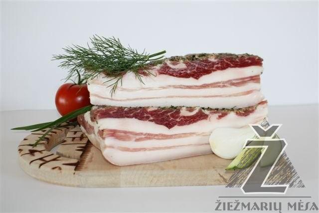 Dwa plastry surowego, peklowanego boczku ułożone na sobie na drewnianej desce z dekoracją z kopru, pomidora i cebuli dymki, znak wodny producenta w prawym dolnym rogu.