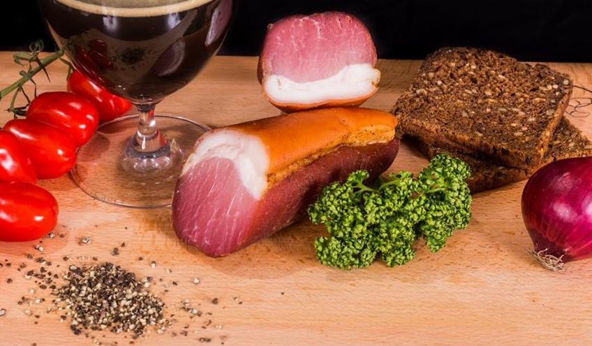 Martwa natura: wędliny z ciemnym piwem, pomidorkami cherry, żytnim chlebem, czerwoną cebulą, brokułami i przyprawami na drewnianej desce.