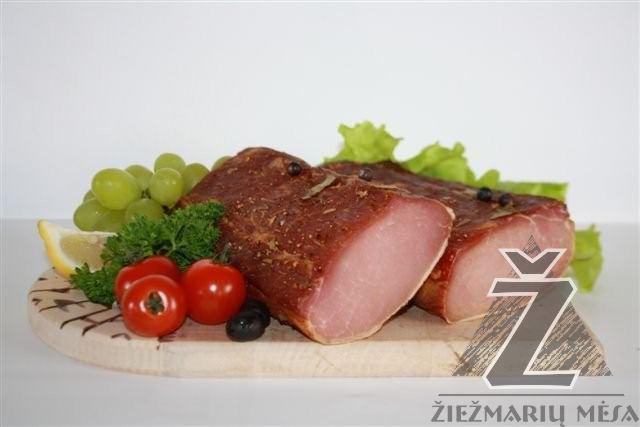 Wędlina w przekroju, udekorowana winogronami, pomidorami, natką pietruszki i cytryną, ułożona na drewnianej desce z widocznym logo producenta w prawym dolnym rogu.