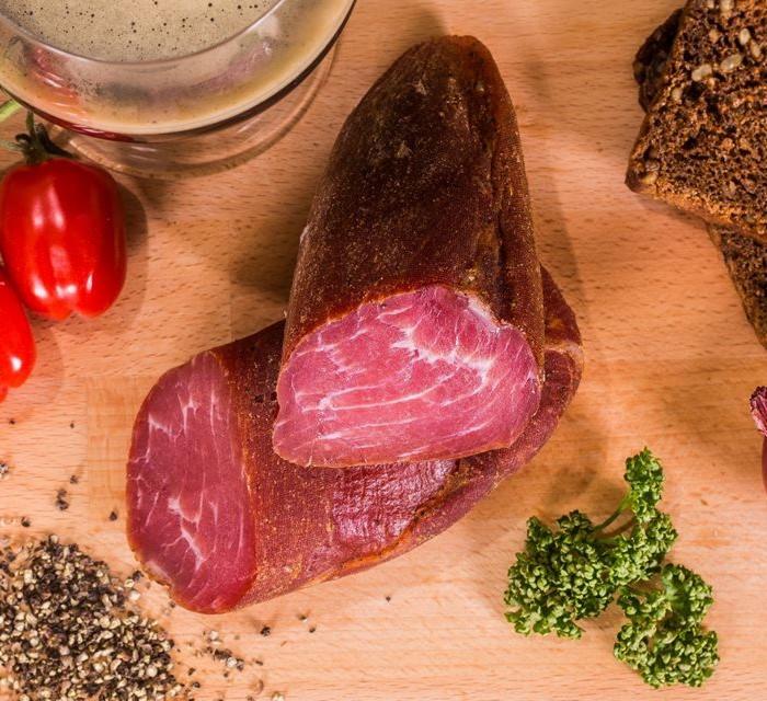 Krojona wędlina z ciemną skórką, ułożona na drewnianej desce z ziarnami pieprzu, pomidorkami koktajlowymi, pieczywem razowym i świeżą natką pietruszki.