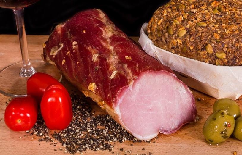 Wędlina wędzona na desce z pomidorami koktajlowymi, oliwkami, pieprzem i chlebem z ziarnami, kieliszek czerwonego wina w tle.