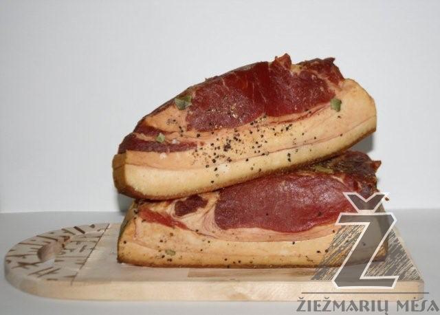 Dwa kawałki wędzonego boczku ułożone jeden na drugim na drewnianej desce do krojenia, posypane pieprzem i ziołami, z widocznym logotypem firmy na desce.