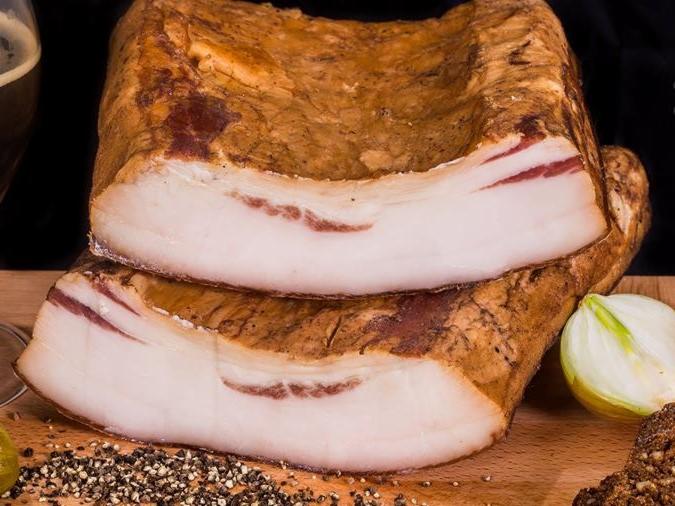 Dwa bloki wędzonego boczku z widocznymi warstwami mięsa i tłuszczu, ułożone na drewnianej desce z czarnym pieprzem w ziarnach, obok cebuli i ciemnego pieczywa, w tle ciemny napój w szklance.