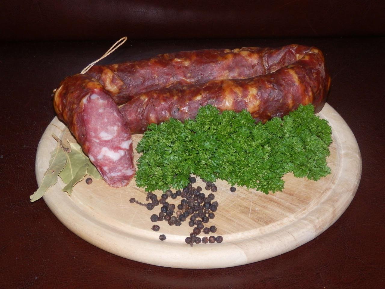 Wędzona kiełbasa zwinięta w okrąg na drewnianej desce, udekorowana świeżą natką pietruszki, ziarnami pieprzu i liściem laurowym.