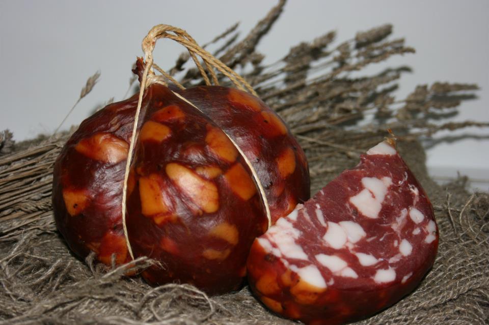 Okrągła wędlina z kawałkami sera, związana sznurkiem, obok plaster wędliny z widocznymi kawałkami tłuszczu, na tle jutowej tkaniny i suszonych traw.