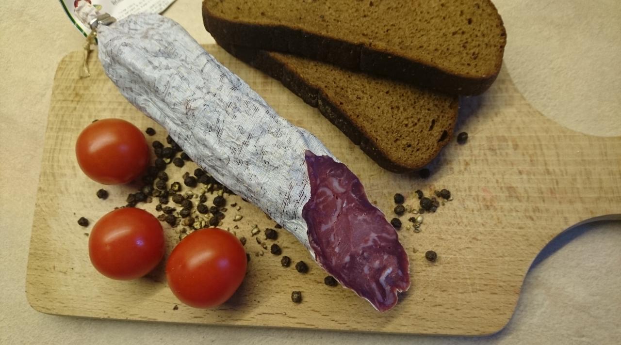 Krojona suszona kiełbasa w białej osłonce, trzy pomidorki koktajlowe, ziarenka pieprzu i dwa kromki ciemnego chleba na drewnianej desce do krojenia.