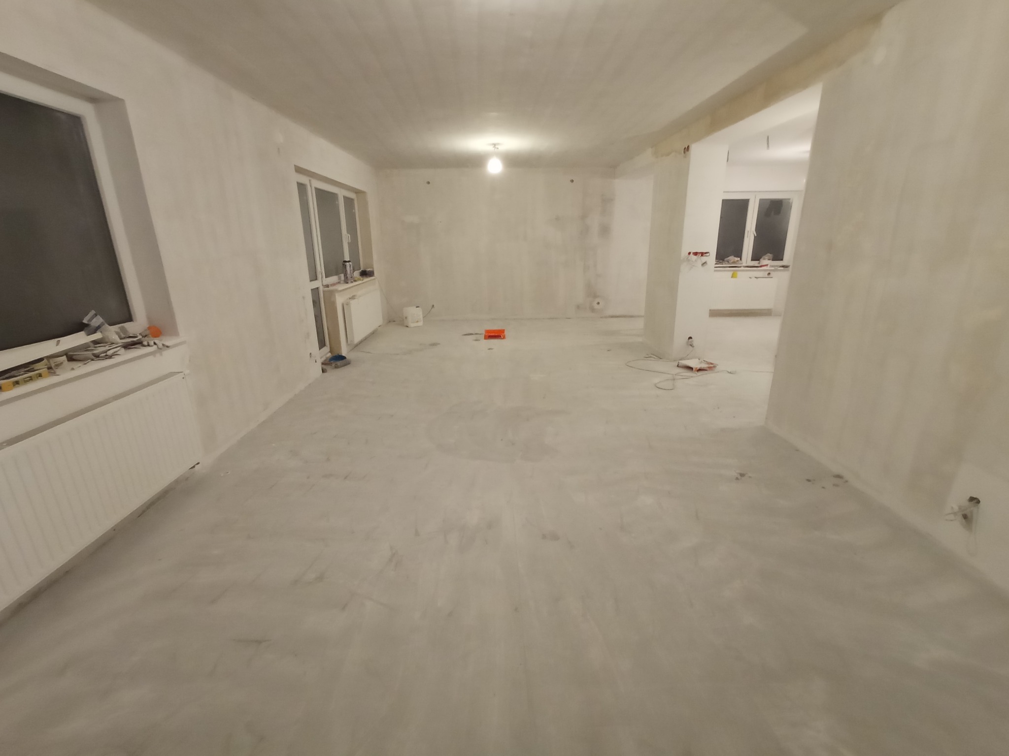 Wnętrze w trakcie szpachlowania ścian i wylewania posadzki, widoczne narzędzia i materiały budowlane, surowy stan wykończenia.