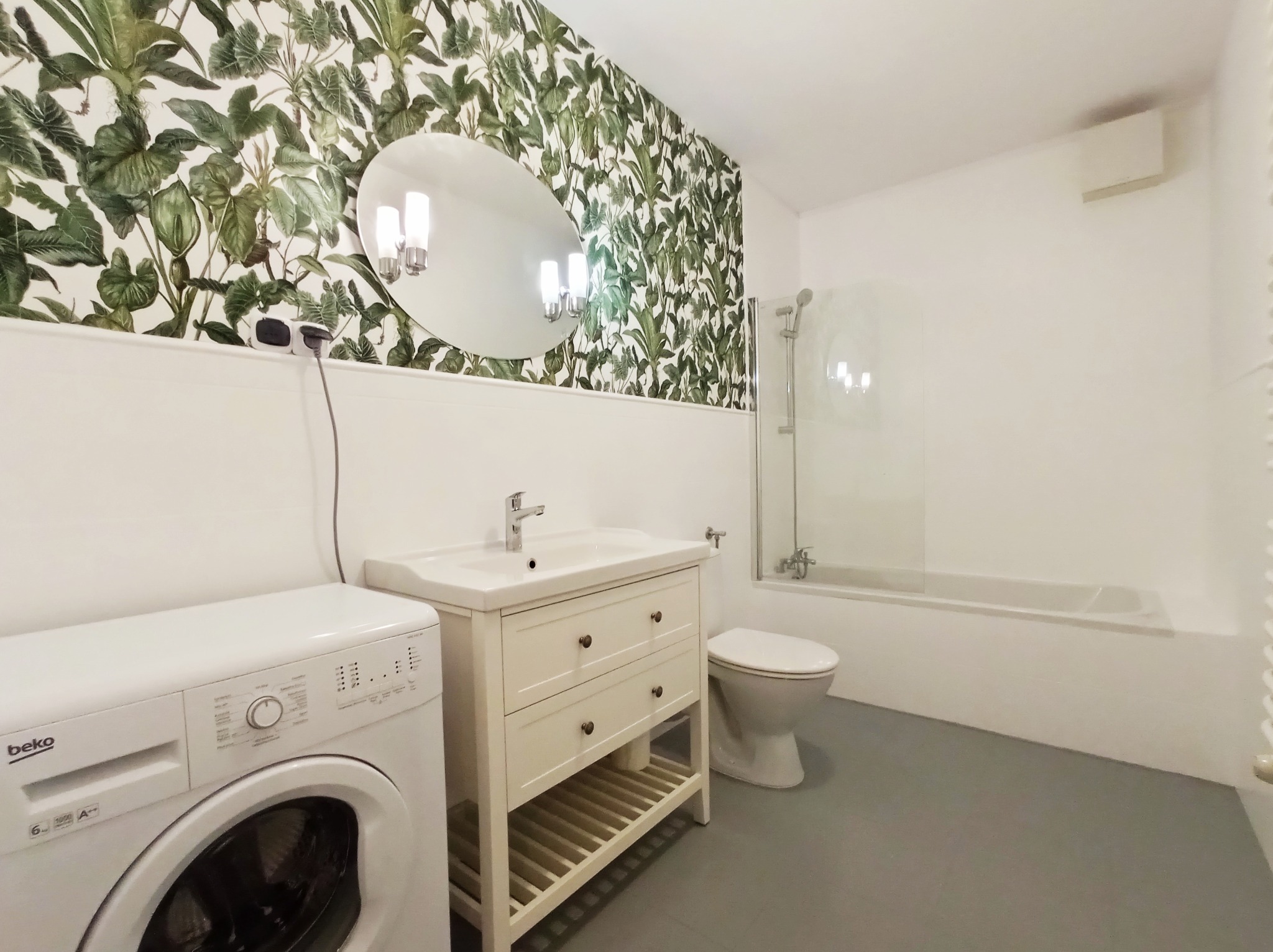 Jasna łazienka z tapetą w motywy roślinne, okrągłe lustro z oświetleniem, biała umywalka z szafką, pralka Beko i wanna za szklanym parawanem.