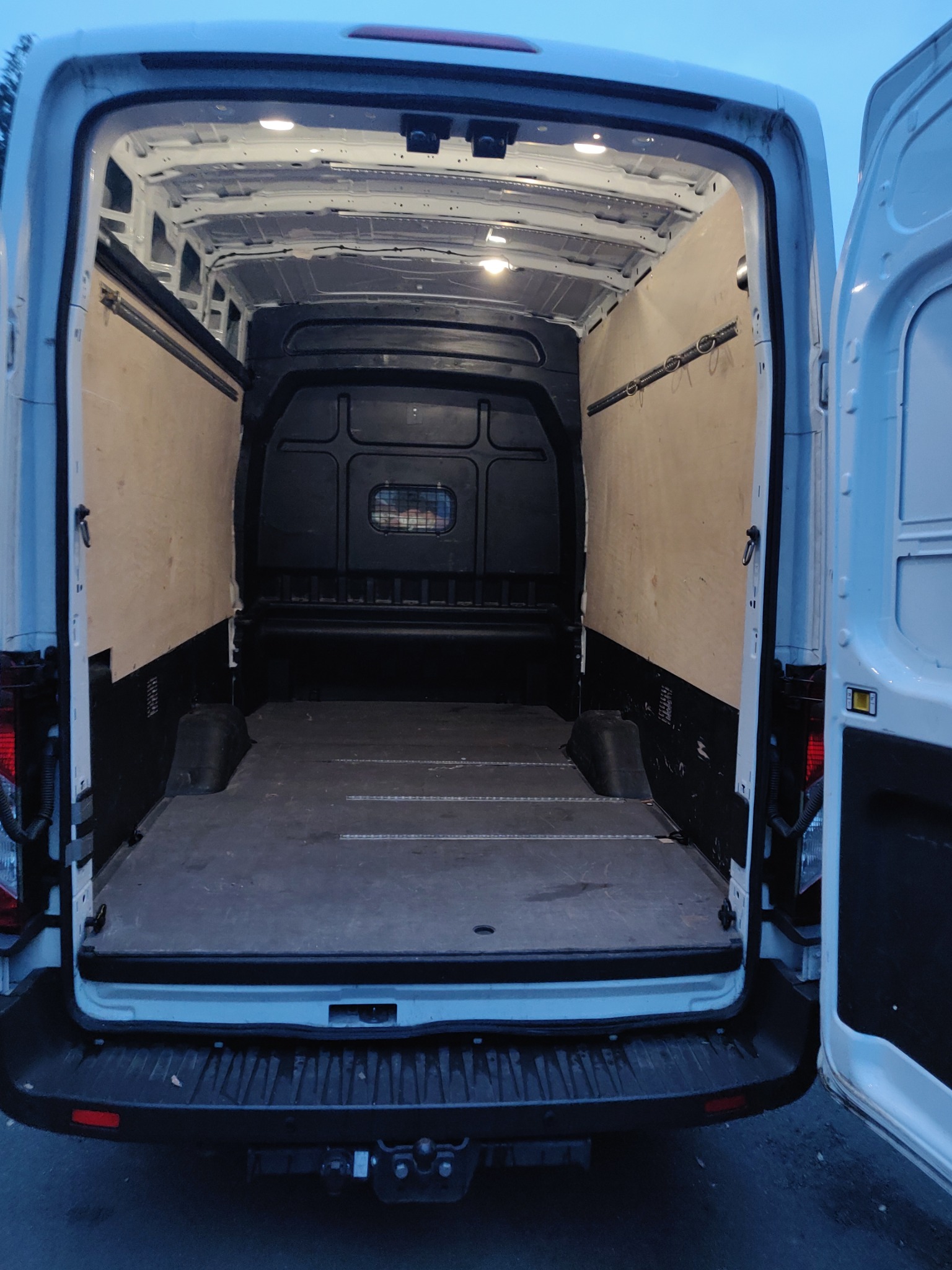 Wnętrze pustej furgonetki z otwartymi tylnymi drzwiami, przygotowanej do transportu, z drewnianą okładziną ścian i oświetleniem sufitowym.