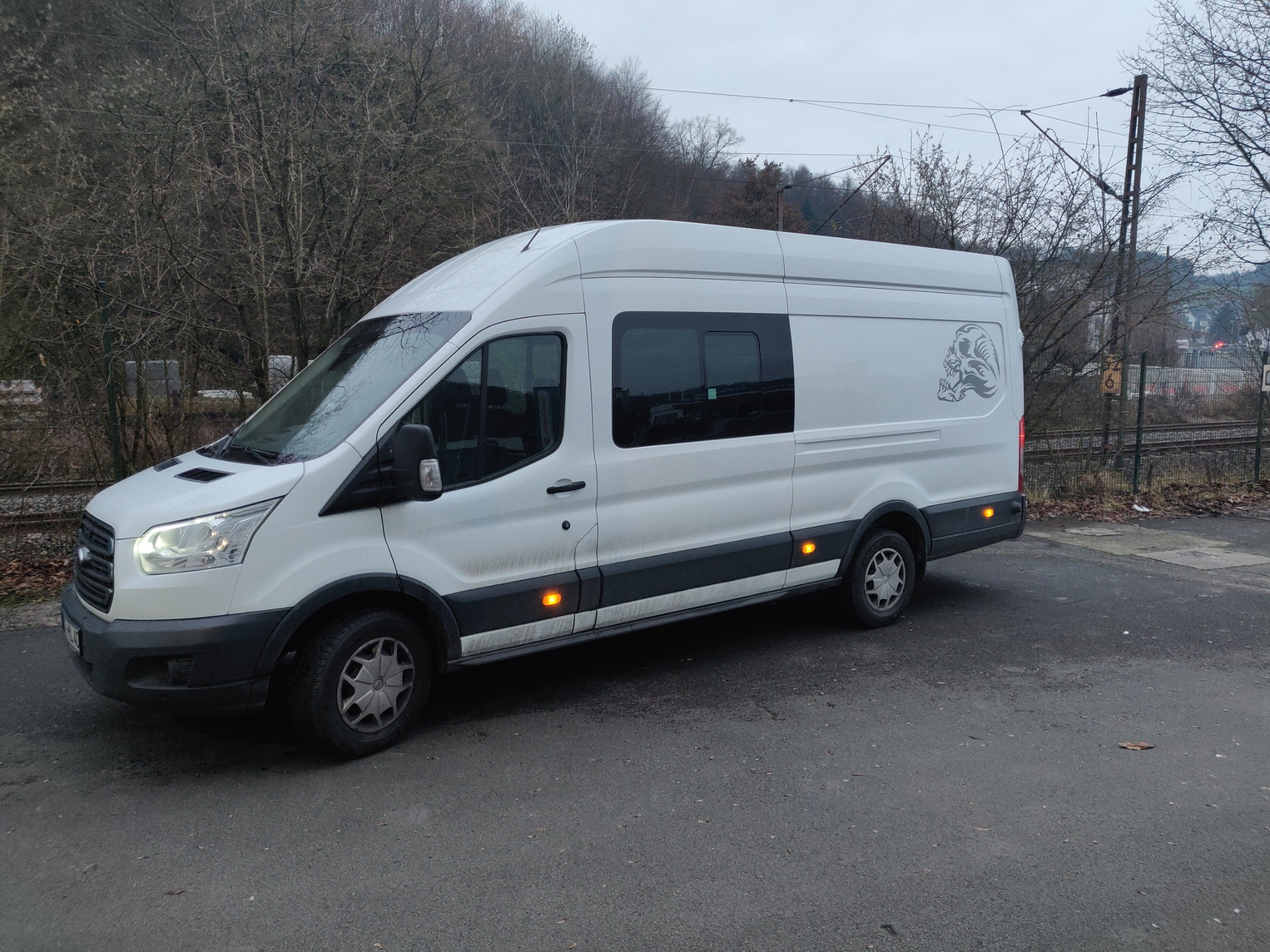 Biały samochód dostawczy z grafiką czaszki na boku, zaparkowany na szarym asfalcie obok torów kolejowych i drzew bez liści.