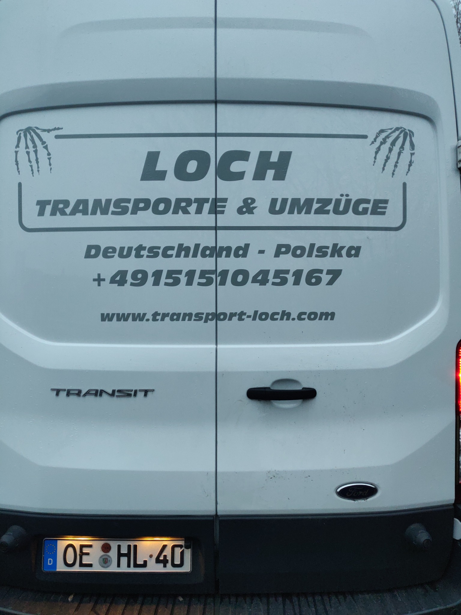 Tył białego furgonetki transportowej firmy Loch Transporte & Umzüge z numerem telefonu, adresem strony internetowej, relacją tras Polska-Niemcy oraz motywem graficznym dłoni szkieletu.