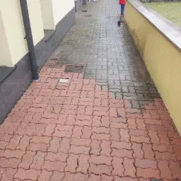 Częściowo umyta czerwona kostka brukowa na posesji, widoczna osoba w czerwonej kurtce z kijem do mycia, kontrast między czystą i brudną powierzchnią.