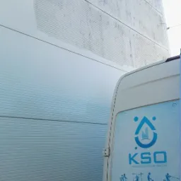 Fragment elewacji hali przemysłowej, z widocznym efektem czyszczenia ciśnieniowego, obok zaparkowany biały bus z logo firmy świadczącej usługi porządkowe.