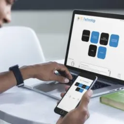 Osoba korzystająca z laptopa i smartfona, na obu urządzeniach wyświetlony interfejs aplikacji z elementami graficznymi w kolorach czarnym i niebieskim, obok leży zielona książka.