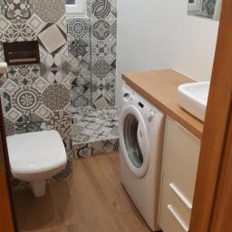 Remonty Wykończenia Wnętrz - Wykończona łazienka z patchworkowymi płytkami w odcieniach szarości i bieli, kabina prysznicowa bez brodzika, toaleta, pralka pod blatem drewnianym z umywalką nablatową, podłoga z paneli...