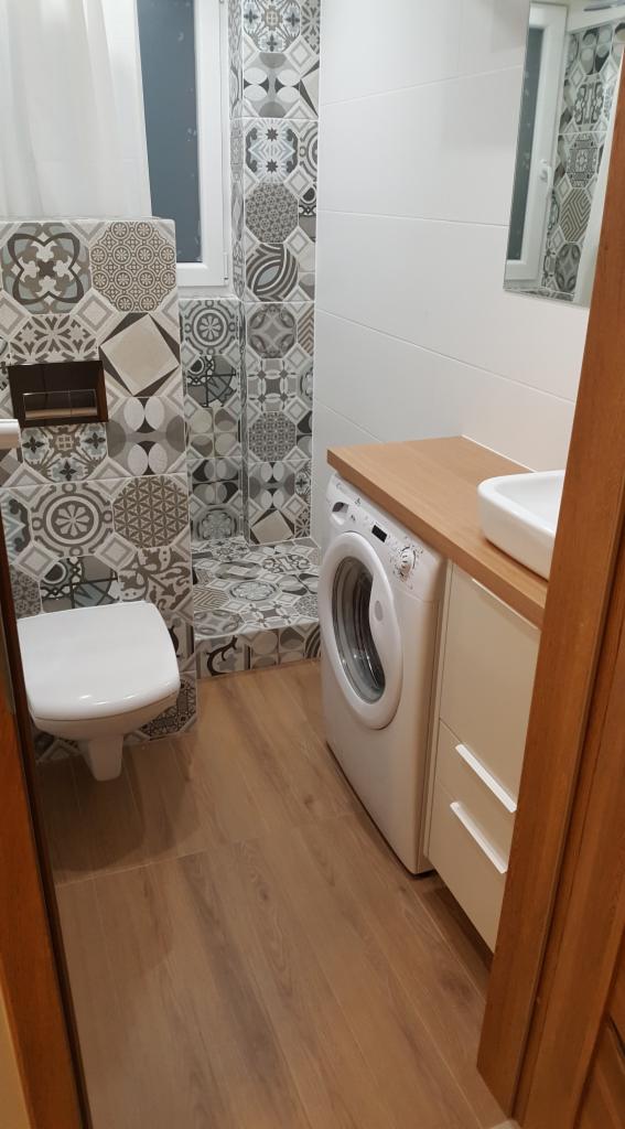 Wykończona łazienka z patchworkowymi płytkami w odcieniach szarości i bieli, kabina prysznicowa bez brodzika, toaleta, pralka pod blatem drewnianym z umywalką nablatową, podłoga z paneli...