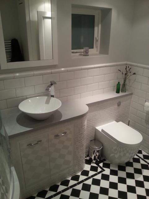 Biała łazienka z umywalką nablatową, szafką w romby, toaletą bezkołnierzową i czarno-białą szachownicą na podłodze; dekoracje: lawenda w wazonie i mała zielona butelka.