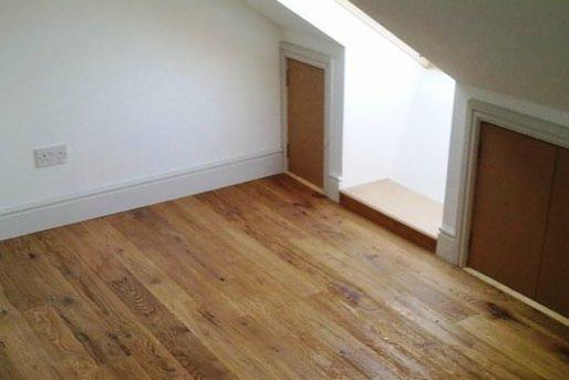 Pokój na poddaszu z drewnianą podłogą, białymi ścianami, listwami przypodłogowymi i zabudowanymi szafkami z drewnianymi drzwiczkami po bokach wnęki.