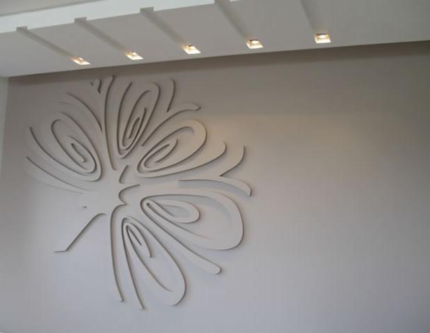 Dekoracyjna, przestrzenna aplikacja ścienna w kształcie stylizowanego kwiatu, wykonana z jasnego materiału, na gładkiej, szarej ścianie w pomieszczeniu z oświetleniem sufitowym.