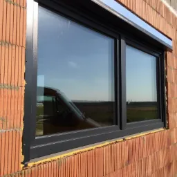 Nowo zamontowane, trzyskrzydłowe okno z szarą ramą w surowej, ceglanej ścianie, z widoczną roletą zewnętrzną i refleksami krajobrazu w szybach.