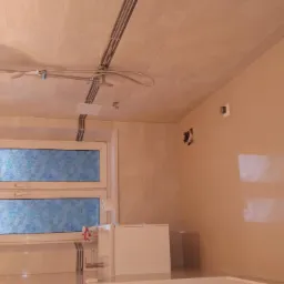 Wykończona łazienka w Budzowie z beżowymi płytkami na ścianach i suficie, widoczna kabina prysznicowa z deszczownicą oraz okno z niebieską folią dekoracyjną.