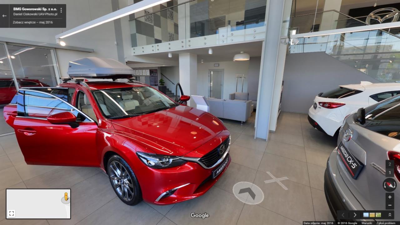 Czerwona Mazda 6 kombi z otwartymi drzwiami w salonie samochodowym, z szarym boxem dachowym, widok na inne modele Mazdy i strefę wypoczynkową.