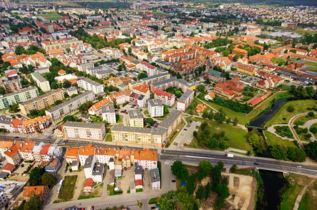 Panoramiczny widok z lotu ptaka na miasto z budynkami mieszkalnymi o zróżnicowanej architekturze, parkiem z rzeką i ruchliwą drogą.