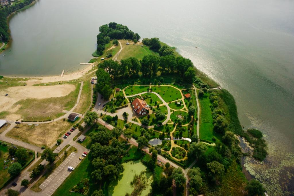 Widok z lotu ptaka na malowniczy półwysep z jeziorem, plażą, budynkiem z czerwonym dachem i starannie utrzymanym parkiem z alejkami.