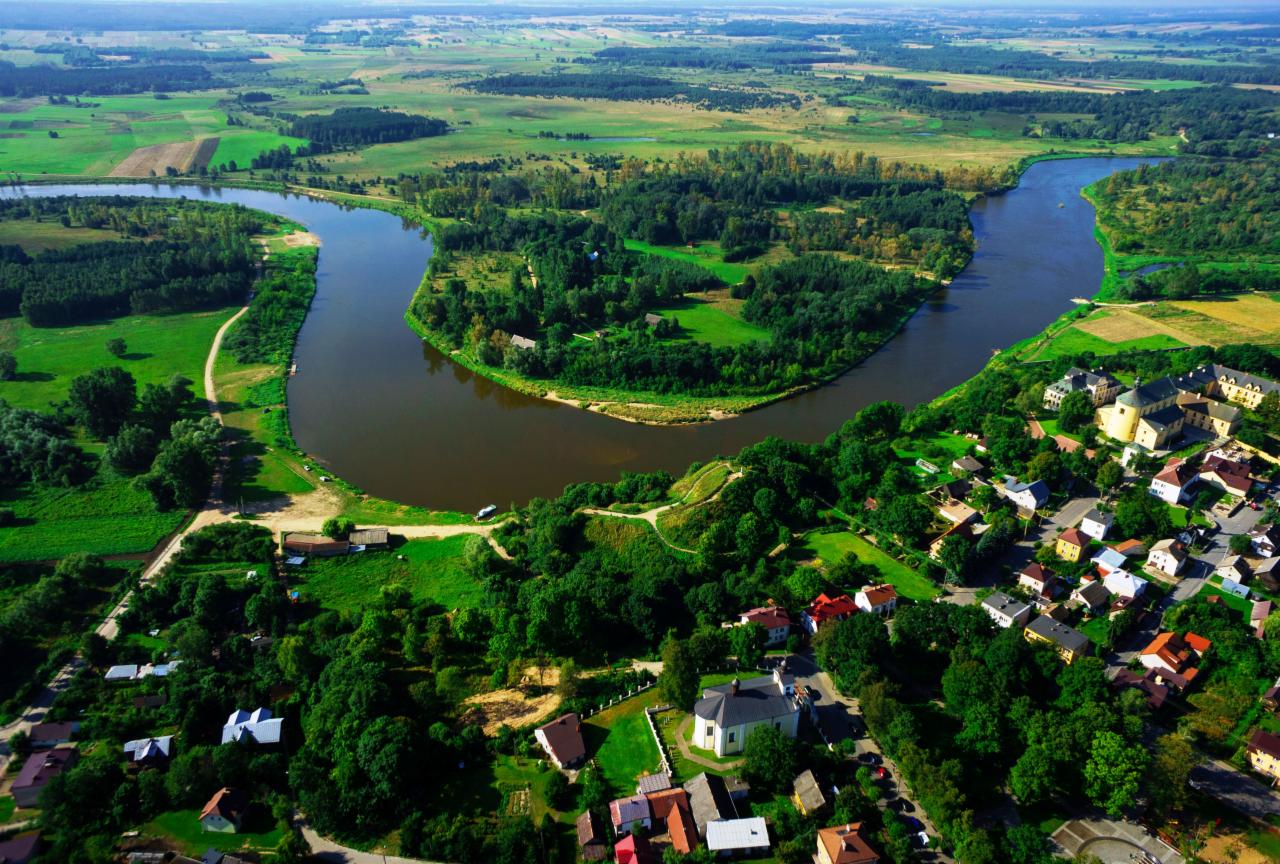 Panoramiczny widok z lotu ptaka na zakole rzeki otoczone zielenią lasów i pól uprawnych, z widoczną zabudową wiejską i kościołem.