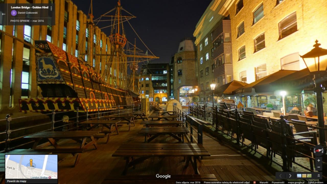 Replika statku Golden Hind zacumowana przy nabrzeżu w Londynie, widok nocny na oświetlony statek z herbem jelenia, stoliki i krzesła kawiarniane na pierwszym planie.